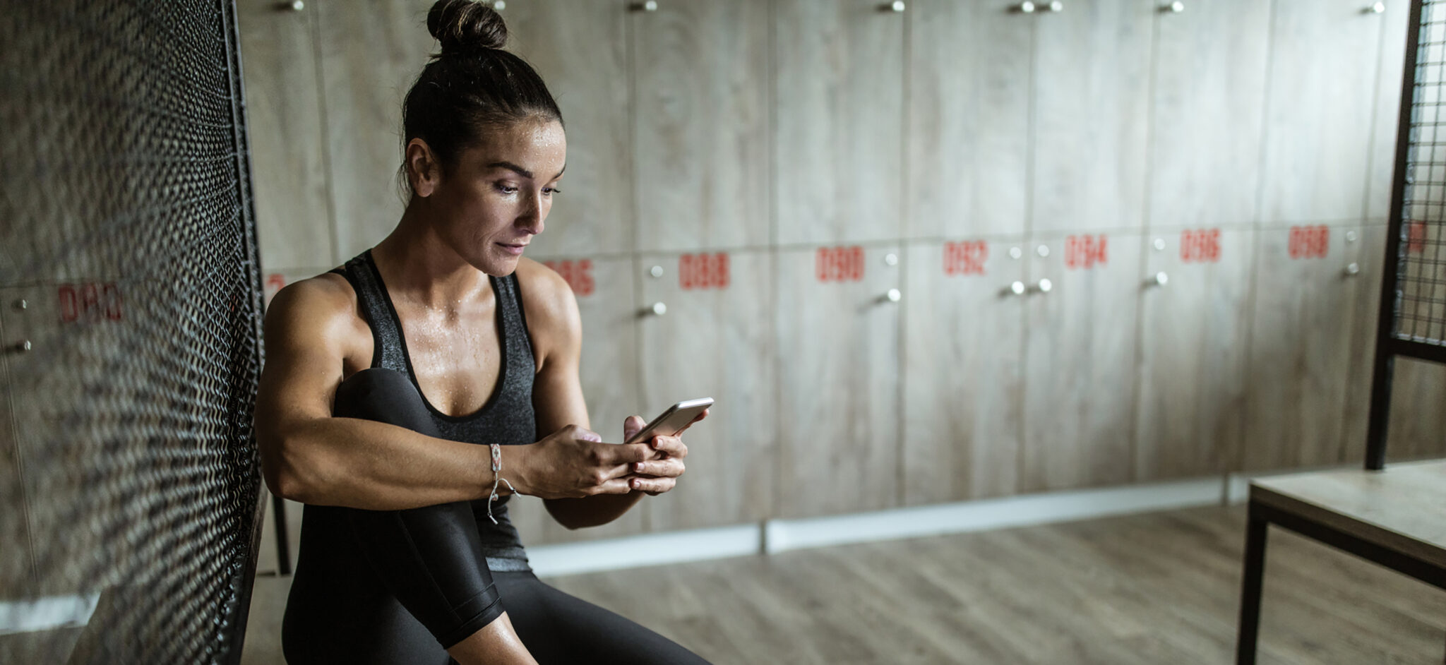 Young athletic woman using mobile phone after sports training at gym's dressing room.