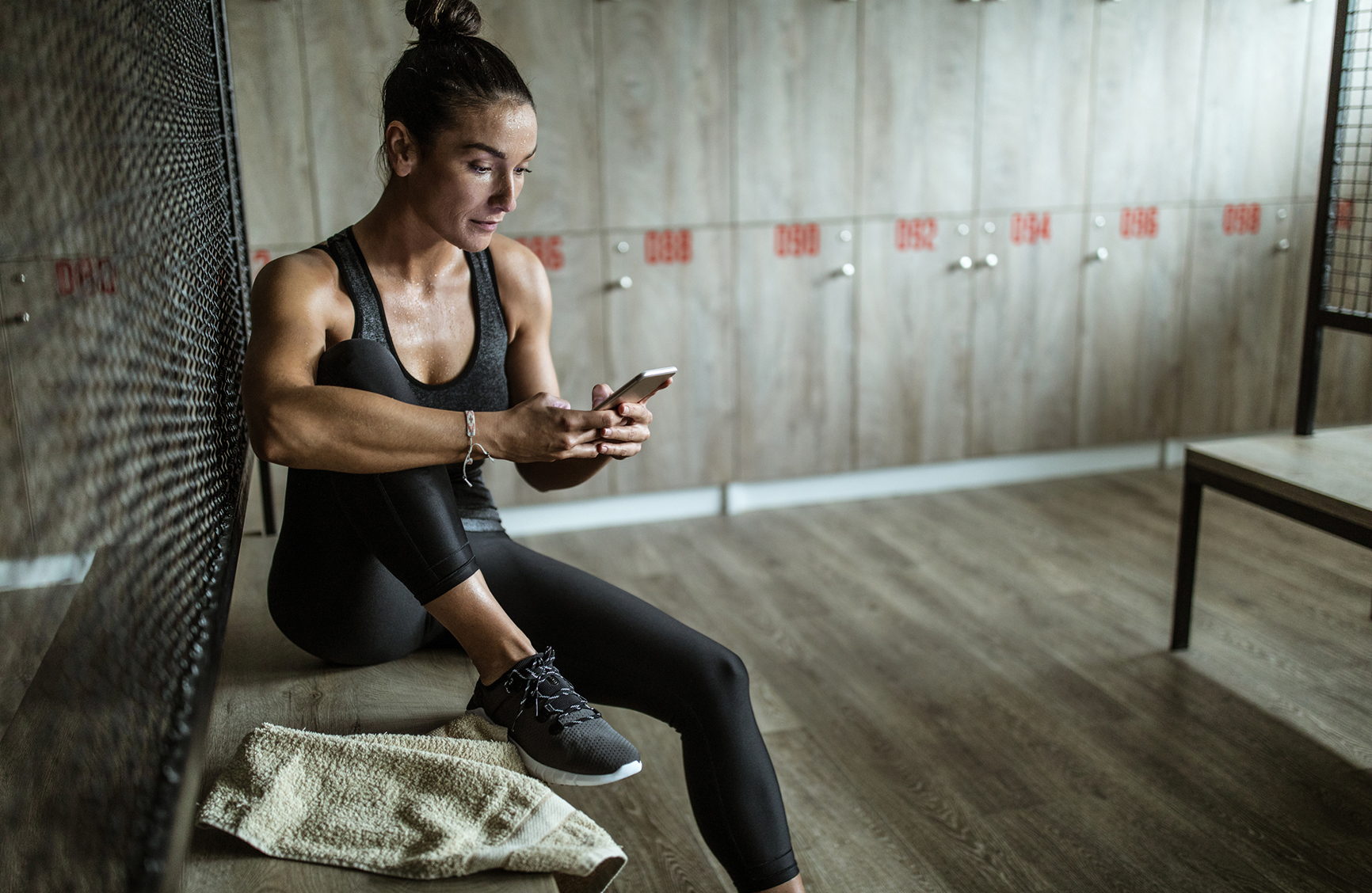 Young athletic woman using mobile phone after sports training at gym's dressing room.