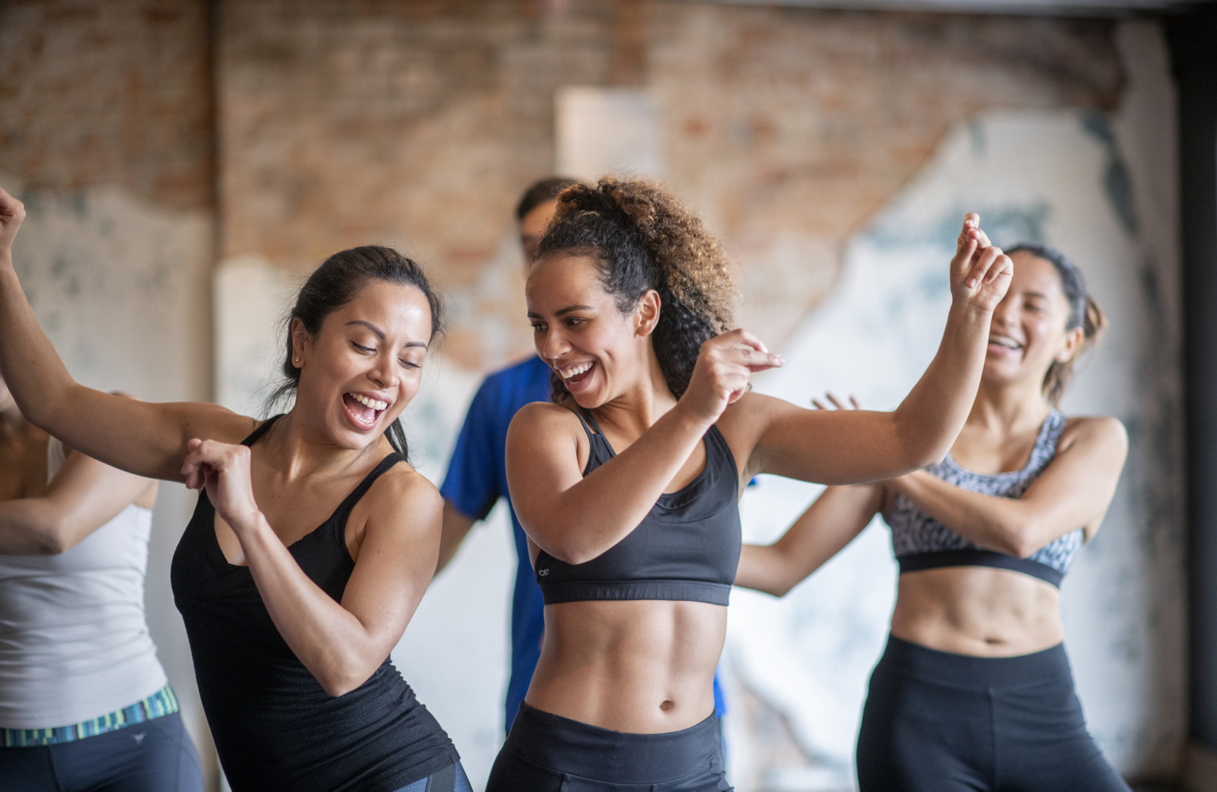 A small group of Hispanic men and women participate in a fitness dance class. They are wearing comfortable athletic wear and have their arms p as they dance and move around the room with joyful expressions. The focus is on two women in front who are bumping hips!