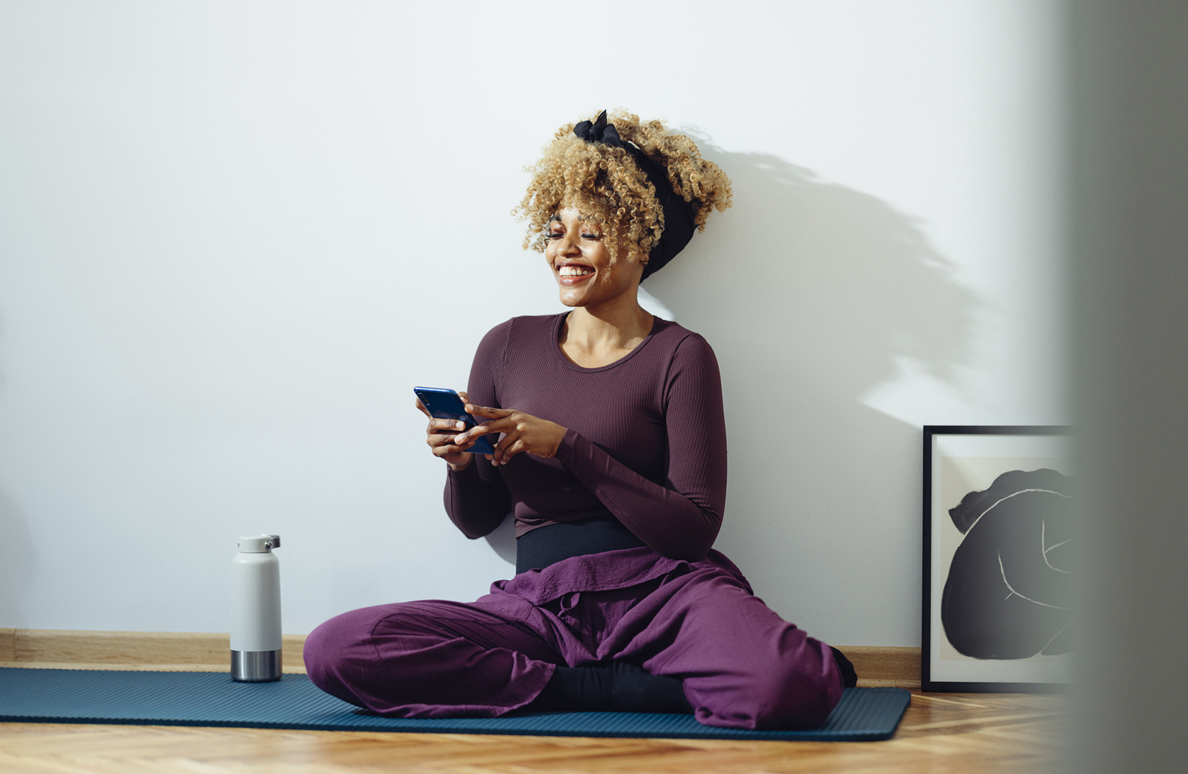 Afro-American woman is sitting on the yoga mat on the floor an holding her phone. She looks happy and trilled. She might be taking a break during an exercise session. She is in a fitness studio or in her room during the day and she might be a yoga student or an instructor. She is laughing.